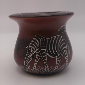 May include: A small, red ceramic pot with a black rim and a white zebra design. The zebra is facing left and is in a walking position.