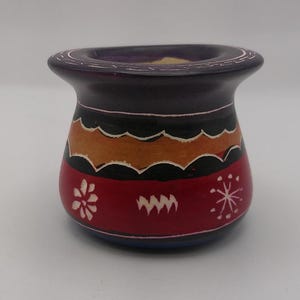 May include: A small, hand-painted ceramic pot with a purple, black, orange, red, and blue design. The pot has a wide mouth and a rounded body.