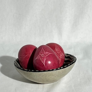 May include: Three pink soapstone eggs with white floral designs are displayed in a white bowl with black stripes.