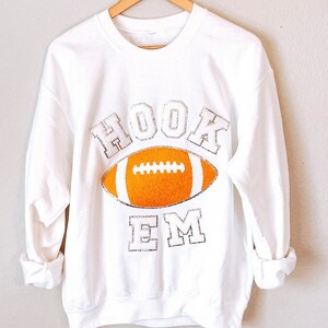 Könnte beinhalten: Weißer Pullover mit einem silbernen Glitzer-Motiv eines Fußballs und den Worten "HOOK EM" auf der Vorderseite.
