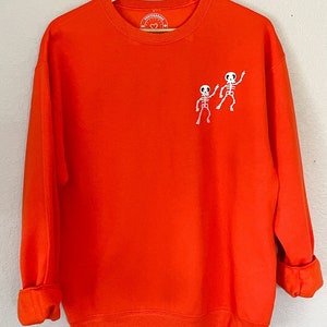 May include: Orange sweatshirt with a small embroidered design of two skeletons dancing on the left chest. The sweatshirt has a crew neck and long sleeves.