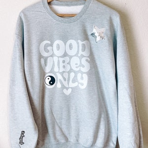 Può includere: Felpa grigia con il testo "GOOD VIBES ONLY" in bianco, un simbolo yin yang e un cuore bianco. La felpa ha una stella scintillante cucita sul lato superiore destro e il testo "feeling it" sul manico sinistro.