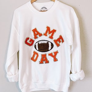 Peut inclure: Sweat-shirt blanc avec un graphisme "GAME DAY" orange et or représentant un ballon de football.