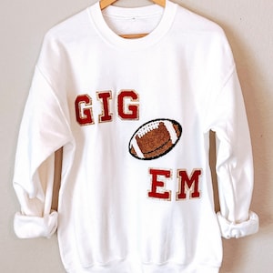 May include: White sweatshirt with the words "GIG 'EM" in red and gold letters with a brown and white football graphic.