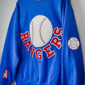 Könnte beinhalten: Ein königsblauer Pullover mit einem roten und weißen Baseball-Motiv und dem Wort "RANGERS" in Rot mit silbernem Glitzer. Der Pullover hat einen roten und weißen Aufnäher auf dem Ärmel, auf dem "Hey Batter Batter Swing!" steht.