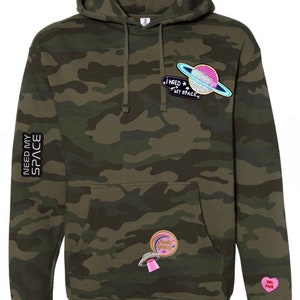 Puede incluir: Una sudadera con capucha de camuflaje verde con una variedad de parches coloridos. Los parches incluyen un planeta con un anillo, un OVNI, un corazón rosa con el texto "Girl Power" y un arcoíris con el texto "Space Galaxy". La sudadera también tiene el texto "I Need My Space" y "Need My Space" en la parte delantera y la manga.