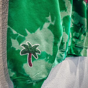 May include: Green tie-dye sweatshirt with a palm tree patch on the sleeve. The sweatshirt has a ribbed cuff and a red and gold lettered design on the upper part of the garment. The fabric has a faded, bleached appearance.