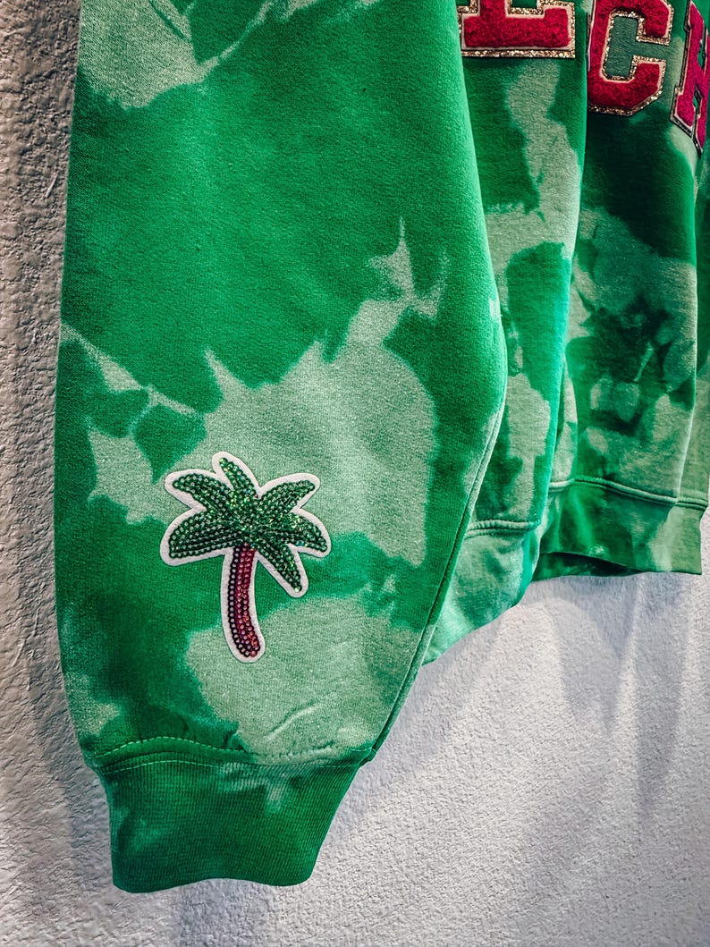 May include: Green tie-dye sweatshirt with a palm tree patch on the sleeve. The sweatshirt has a ribbed cuff and a red and gold lettered design on the upper part of the garment. The fabric has a faded, bleached appearance.