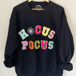 Pode incluir: Camisola de moletom preta com um gráfico colorido de "Hocus Pocus" que inclui uma teia de aranha. As letras são delineadas com glitter dourado.