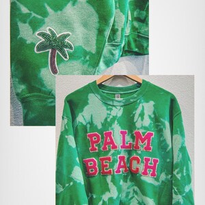 May include: Green tie-dye sweatshirt with the words "PALM BEACH" in pink block letters. A palm tree patch is on the sleeve. The sweatshirt is hanging on a wooden hanger.