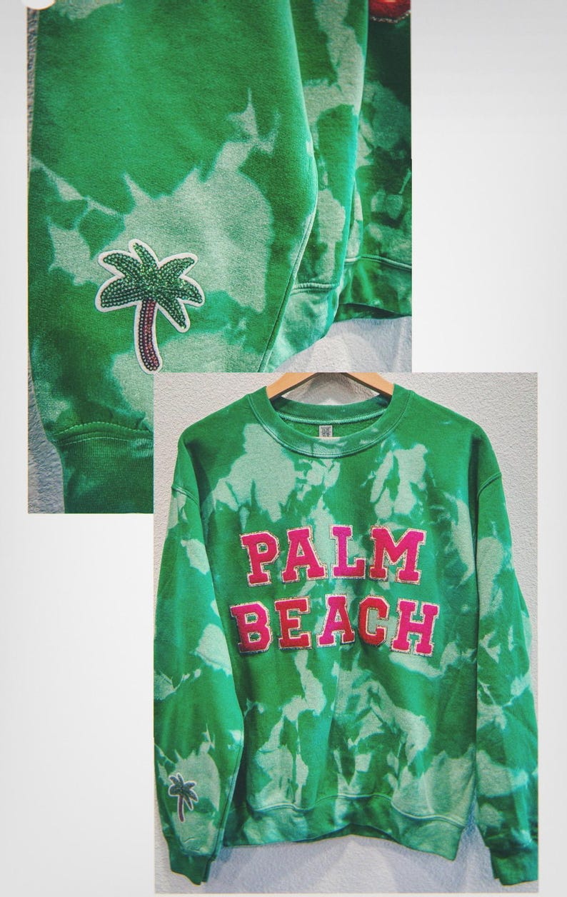 May include: Green tie-dye sweatshirt with the words "PALM BEACH" in pink block letters. A palm tree patch is on the sleeve. The sweatshirt is hanging on a wooden hanger.