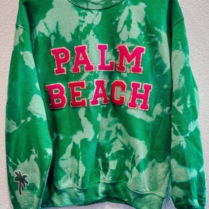 Puede incluir: Sudadera verde teñida con la palabra "PALM BEACH" en letras grandes y rosas. Una pequeña palmera bordada está en la manga izquierda. La sudadera está colgada en una percha de madera.
