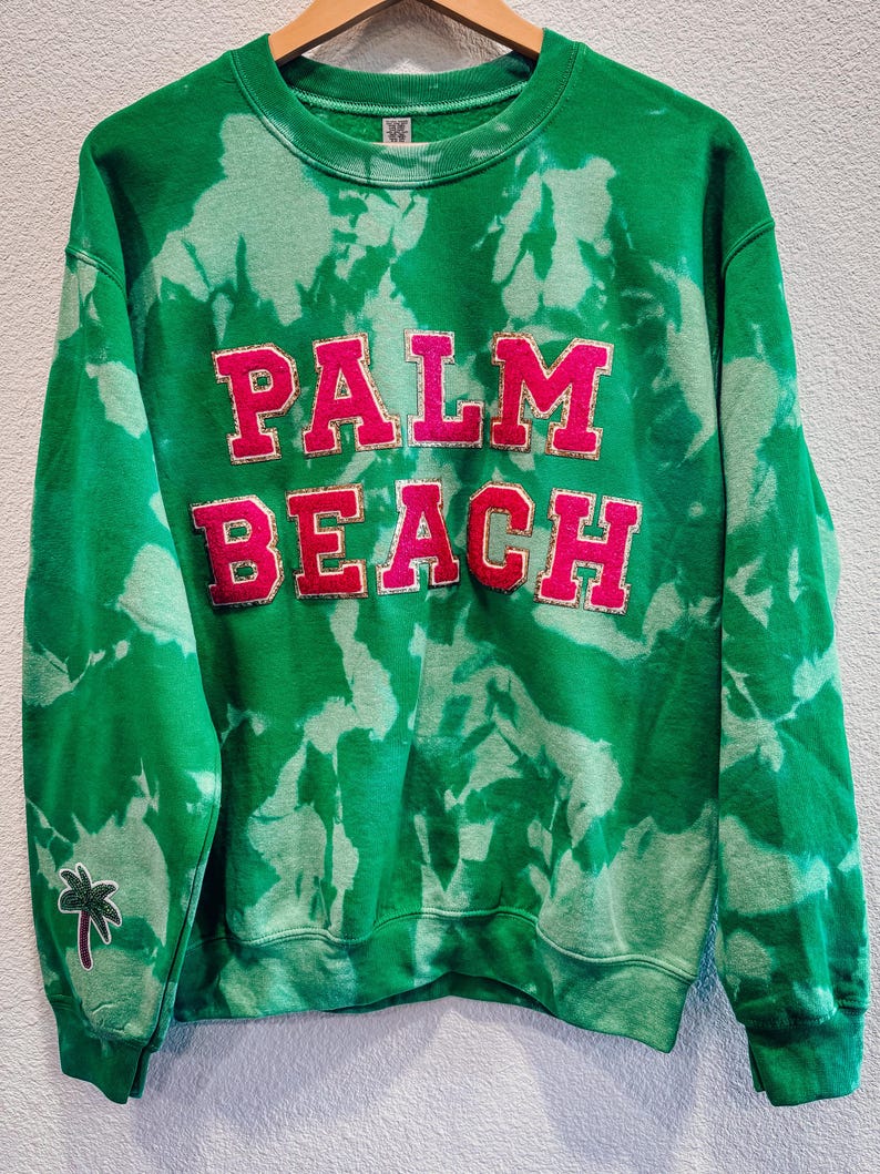 May include: Green tie-dye sweatshirt with the words "PALM BEACH" in large, pink, block letters. A small embroidered palm tree is on the left sleeve. The sweatshirt is hanging on a wooden hanger.