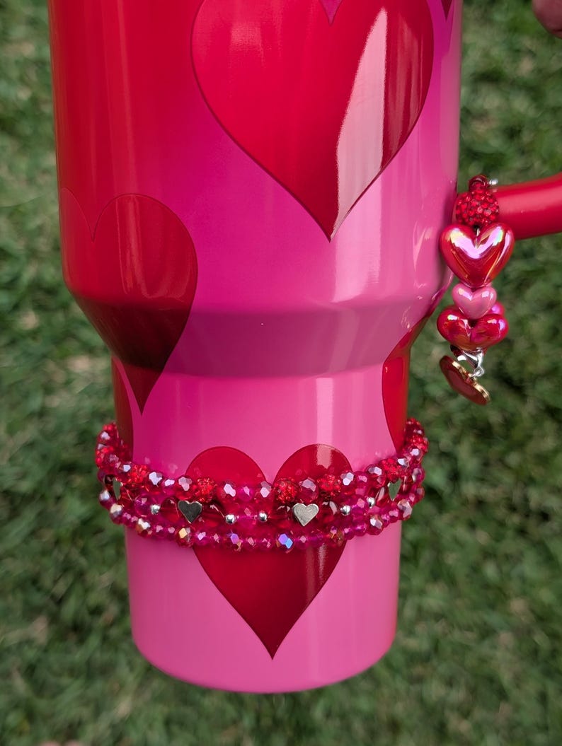 May include: A red and pink tumbler with a heart design. The tumbler features a beaded bracelet and a charm with heart-shaped beads. The handle is red. The tumbler has a Valentine's Day theme.
