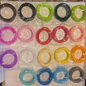 Puede incluir: Una colección de veinte anillos protectores circulares de silicona en varios colores sólidos, incluyendo marrón, negro, verde lima, verde azulado oscuro, verde brillante, magenta, rosa, rojo, coral, rosa claro, morado, blanco, crema, amarillo, naranja, azul claro, azul y gris.