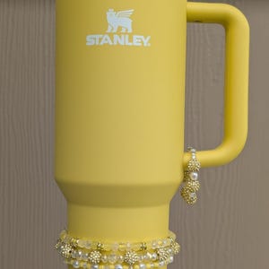 Op de afbeelding: Een gele Stanley-beker met een handvat, een wit deksel en het Stanley-logo. De beker is versierd met twee armbanden met gouden en parel kralen. De armbanden zijn om de beker gewikkeld.