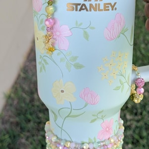 Puede incluir: Vaso Stanley azul claro adornado con motivos florales rosas y amarillos. El vaso presenta un dije de cuentas en el asa y una banda de cuentas alrededor de la base. El logotipo de Stanley es visible en la parte delantera.