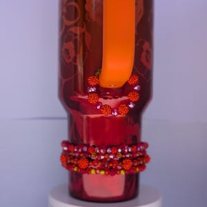 Peut inclure: Un gobelet rouge avec une poignée orange et un motif floral. Le gobelet est orné d'ornements de perles orange et rouges. La poignée est de couleur orange vif et le gobelet repose sur une surface blanche.