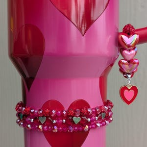 May include: A pink tumbler with red heart designs and a beaded bracelet. The tumbler has a handle with a charm of three iridescent hearts and a heart pendant. The bracelet is red with silver heart accents.