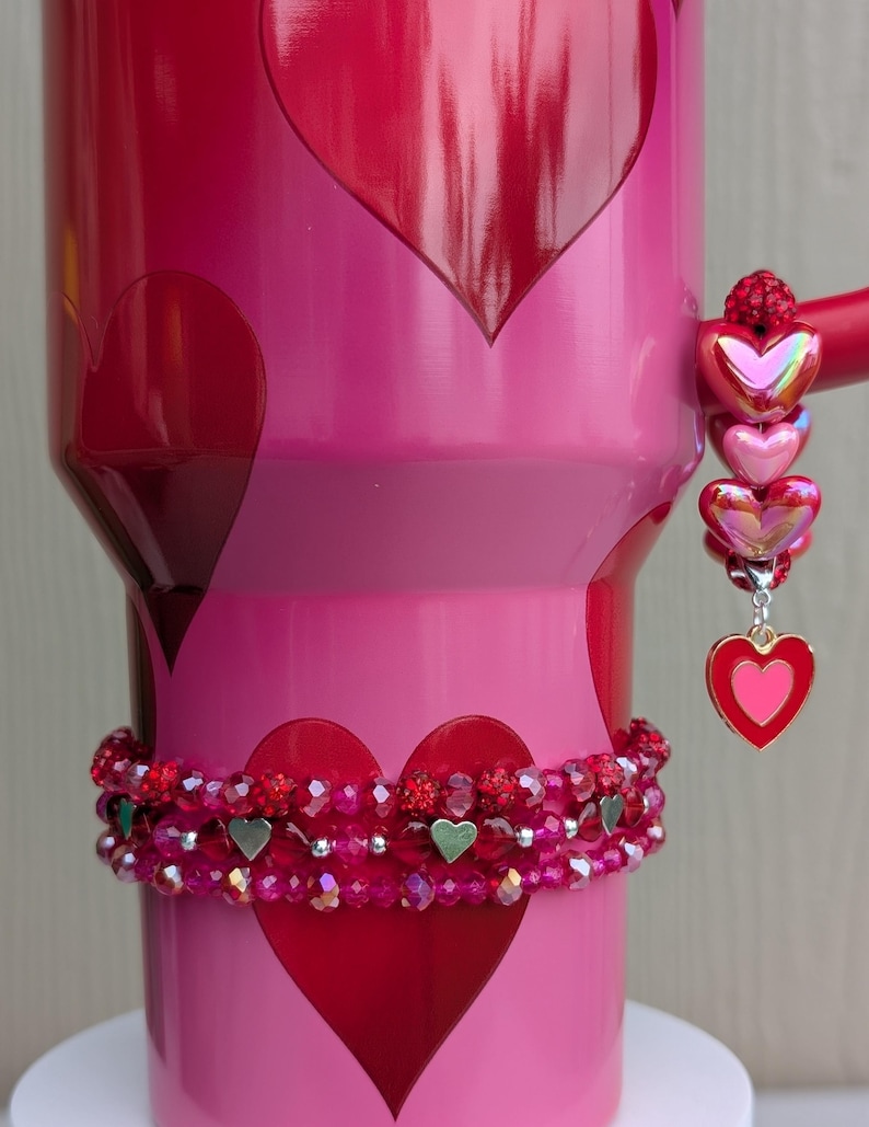 May include: A pink tumbler with red heart designs and a beaded bracelet. The tumbler has a handle with a charm of three iridescent hearts and a heart pendant. The bracelet is red with silver heart accents.