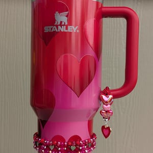 May include: A red and pink Stanley tumbler with a handle, decorated with heart shapes. A beaded bracelet and a charm with heart pendants adorn the tumbler. The Stanley logo is visible on the side.
