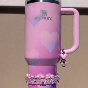 May include: Pink Stanley tumbler with a handle, decorated with heart patterns. The tumbler has a decorative beaded bracelet around the base and a charm on the handle. The Stanley logo is visible on the side.