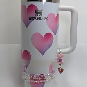 Könnte beinhalten: Weißer Stanley-Becher mit Griff, verziert mit rosa und lila Herzformen. Der Becher hat einen transparenten Deckel und einen Strohhalm. Ein Perlenarmband und ein herzförmiger Anhänger hängen am Griff. Das Stanley-Logo ist sichtbar.