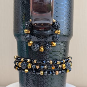 May include: A black tumbler with a snakeskin texture, adorned with two beaded bracelets. The bracelets feature black and gold beads, adding a touch of glamour. The tumbler is set on a white surface.
