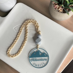 Pode incluir: Um pendente de carro em acrílico transparente com um design de montanha turquesa e as palavras "Adventure Awaits". Ele pende de um cordão trançado dourado e apresenta contas cinza claro. O pendente repousa sobre uma bandeja branca.