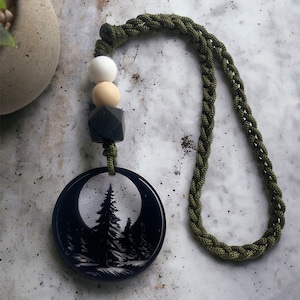 May include: A black pendant necklace featuring a forest scene within a crescent moon shape. The necklace has a braided olive green cord and is accented with white, tan, and black beads. The pendant is approximately 2 inches in diameter.