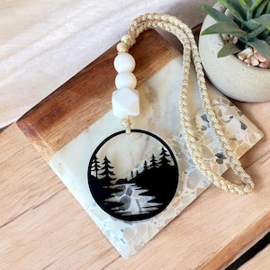 May include: A black circular pendant featuring a forest and river scene, suspended from a beige braided cord. The pendant is adorned with white beads and a geometric shape. The necklace rests on a wooden and marble surface.