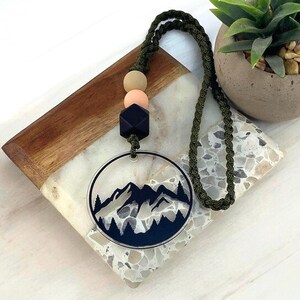 May include: A clear acrylic pendant with a mountain and forest design, suspended from a dark green braided cord. The pendant is accented with wooden beads and a black geometric bead. The necklace rests on a decorative surface.