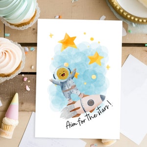 Może przedstawiać: Biała kartka z akwarelową ilustracją astronauty w rakiecie lecącej przez niebieską chmurę z żółtymi gwiazdami. Pod rakietą widnieje napis "Celuj w gwiazdy!"