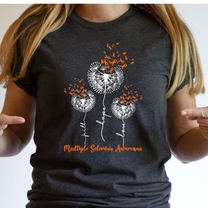 Puede incluir: Una camiseta gris oscuro con un diseño de tres dientes de león blancos con cintas naranjas que se van volando con el viento. Los dientes de león están etiquetados con las palabras "fe", "esperanza" y "amor". El texto "Concientización sobre la esclerosis múltiple" está impreso debajo de los dientes de león.
