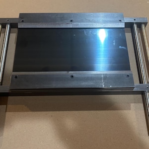 May include: A metal and steel tool with a rectangular black plate in the center. The tool has two horizontal metal bars with a ruler etched on the top bar. Four vertical metal rods support the structure.