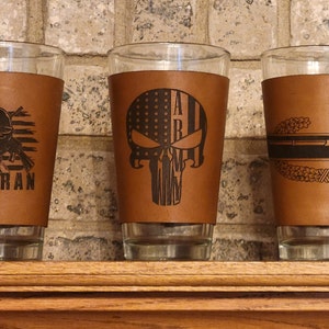 Könnte beinhalten: Drei braune Leder-Bierkühler mit verschiedenen Designs. Der erste Bierkühler zeigt einen Soldaten mit einer Waffe und das Wort "Veteran". Der zweite Bierkühler zeigt einen Schädel mit einer amerikanischen Flagge und das Wort "Army". Der dritte Bierkühler zeigt ein militärisches Abzeichen mit einem Gewehr und einem Kranz.