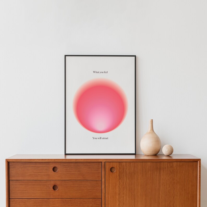 May include: A framed print with a pink gradient circle and the text "What you feel, You will attract". The print is hanging on a white wall above a wooden dresser.
