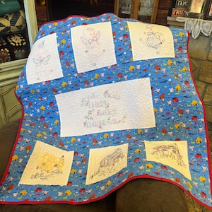 May include: A handmade quilt featuring embroidered images of pigs and houses on a blue background. The quilt has a red border and includes the text "ONCE AGO THERE WERE THREE PIGS". The quilt is a charming, handcrafted item.
