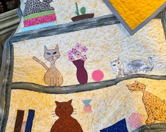 May include: A handmade quilt featuring various cat appliqués. The quilt showcases a yellow background with multiple shelves, each adorned with cat designs, flowers, and decorative elements. The quilt's border is gray, and the reverse side is bright yellow.