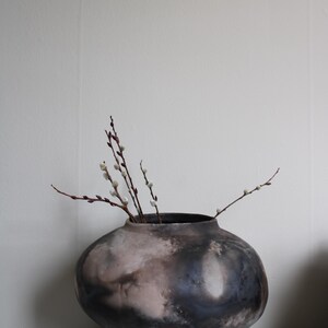 Peut inclure: Un grand vase en céramique fait à la main avec une finition sombre et mouchetée, contenant plusieurs branches de chatons. Le vase a un corps arrondi et un col étroit. La palette de couleurs comprend des nuances de gris, de brun et de crème.