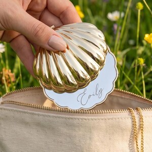 May include: A gold-toned, shell-shaped compact mirror with the name Emily inscribed in cursive script, held above a beige canvas zippered pouch featuring a gold sun charm attached to the zipper pull, set against a backdrop of green grass.