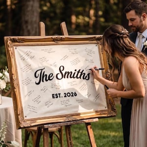 May include: A rectangular white fabric guest book sign with black text reading The Smiths and EST. 2026, displayed in an ornate gold vintage-style frame on a wooden easel. The fabric features handwritten signatures and messages in black ink.