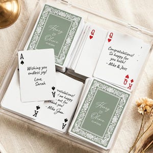 May include: A clear acrylic box containing custom playing cards with sage green backs featuring white filigree borders and the text Henry & Olivia 12.09.2026. Several cards are face up, displaying handwritten-style congratulatory messages.