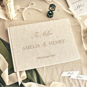 K&ouml;nnte beinhalten: Ein cremefarbenes Hochzeitsg&auml;stebuch mit den Namen "AMELIA & HENRY" und dem Datum "10th January 2026" in Gold. Das Bild zeigt Hochzeitsringe, Schreibwaren und Blumenschmuck, was eine romantische Szene ergibt.