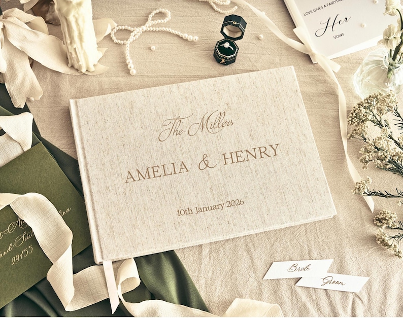 K&ouml;nnte beinhalten: Ein cremefarbenes Hochzeitsg&auml;stebuch mit den Namen "AMELIA & HENRY" und dem Datum "10th January 2026" in Gold. Das Bild zeigt Hochzeitsringe, Schreibwaren und Blumenschmuck, was eine romantische Szene ergibt.
