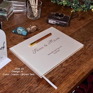 K&ouml;nnte beinhalten: Cremefarbenes G&auml;stebuch im A4-Format mit braunem Text. Auf dem Einband steht "THOMAS & MELANIE" und "Newcastle Upon Tyne, UK 02.08.2025". Ein Holzstift mit goldener Spitze liegt auf dem Buch. Ein wei&szlig;es Lesezeichen ist sichtbar.