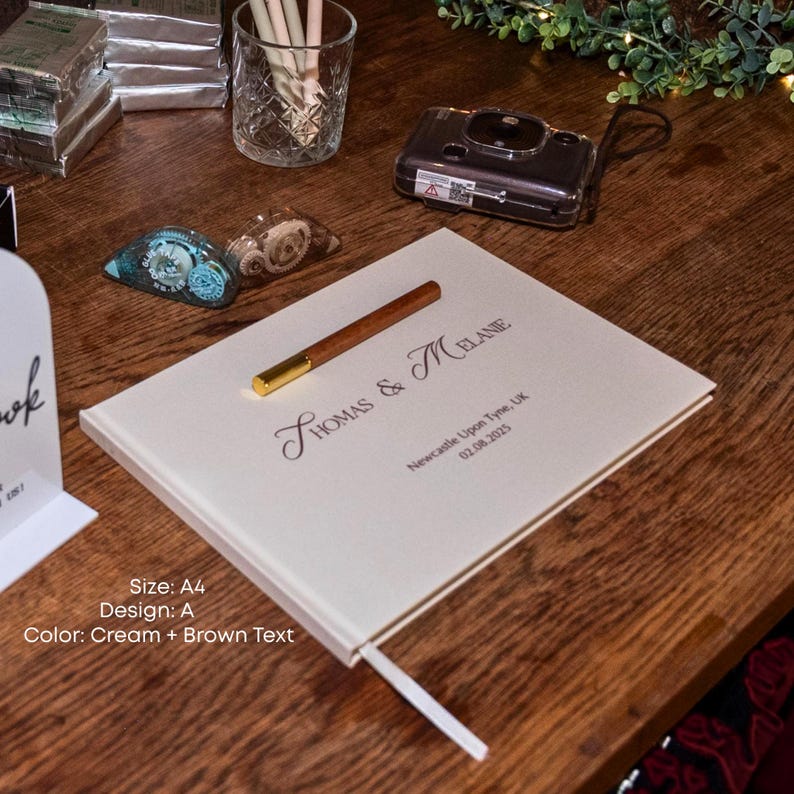 K&ouml;nnte beinhalten: Cremefarbenes G&auml;stebuch im A4-Format mit braunem Text. Auf dem Einband steht "THOMAS & MELANIE" und "Newcastle Upon Tyne, UK 02.08.2025". Ein Holzstift mit goldener Spitze liegt auf dem Buch. Ein wei&szlig;es Lesezeichen ist sichtbar.