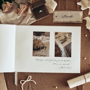 K&ouml;nnte beinhalten: Ein offenes Hochzeitsfotoalbum mit zwei gerahmten Bildern einer Hochzeitstafel. Ein Diamantring in einer braunen Samtbox, ein Etikett mit der Aufschrift "Bride" und eine Schriftrolle sind ebenfalls zu sehen. Das Album enth&auml;lt das Zitat "Was Gott zusammengef&uuml;gt hat, soll der Mensch nicht trennen."