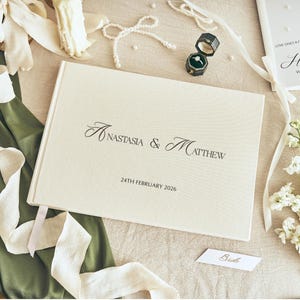 K&ouml;nnte beinhalten: Eine Hochzeitskomposition mit einem cremefarbenen G&auml;stebuch mit den Namen "Anastasia & Matthew" und dem Datum "24. Februar 2026". Die Szene umfasst Eheringe, Blumen und B&auml;nder, die eine romantische Atmosph&auml;re schaffen.