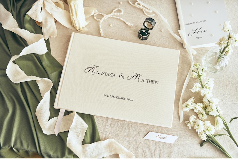 K&ouml;nnte beinhalten: Eine Hochzeitskomposition mit einem cremefarbenen G&auml;stebuch mit den Namen "Anastasia & Matthew" und dem Datum "24. Februar 2026". Die Szene umfasst Eheringe, Blumen und B&auml;nder, die eine romantische Atmosph&auml;re schaffen.